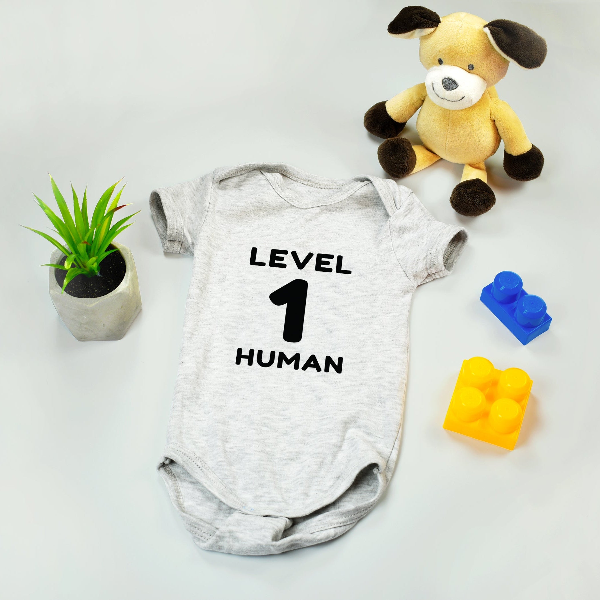 Baby onesie with 'Level 1 Human' text, plush toy, and colorful blocks on a light background