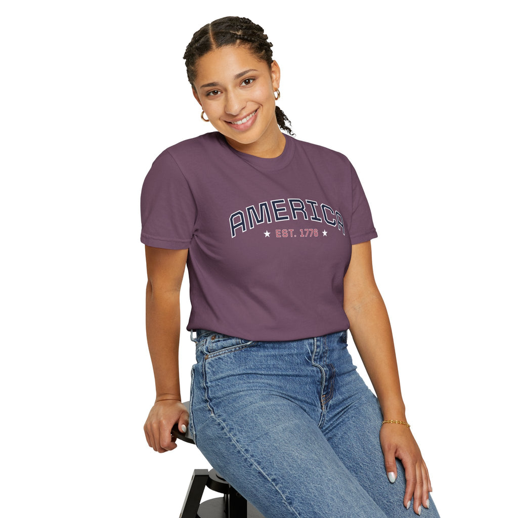 a woman wearing a berry t-shirt with 'AMERICA EST. 1776' typography design in varsity style on a white background