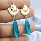 Gold earrings with blue teardrop-shaped stones held between fingers against a neutral background
