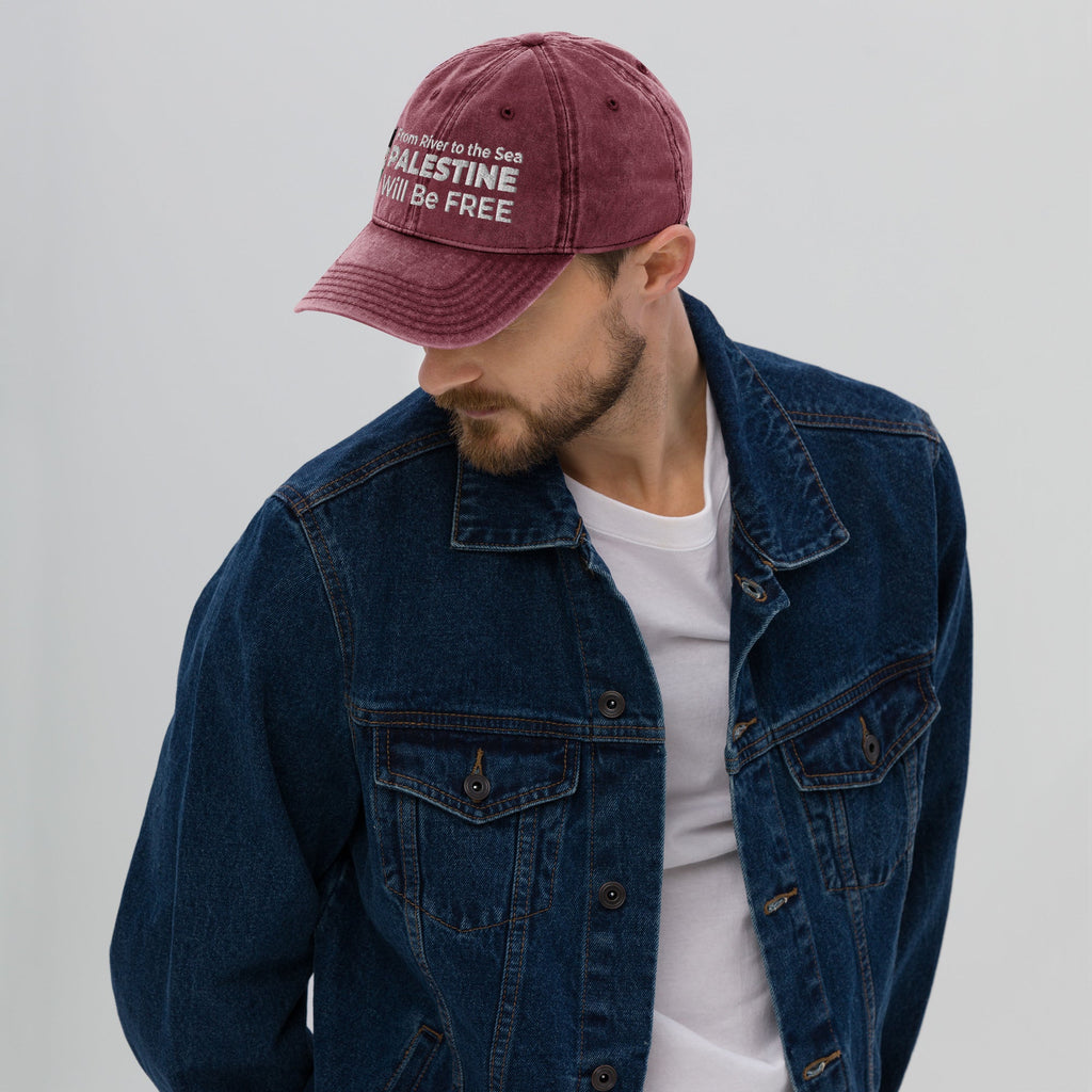 A man wearing a denim jacket and a maroon cap with the text and Palestine flag, on a plain background