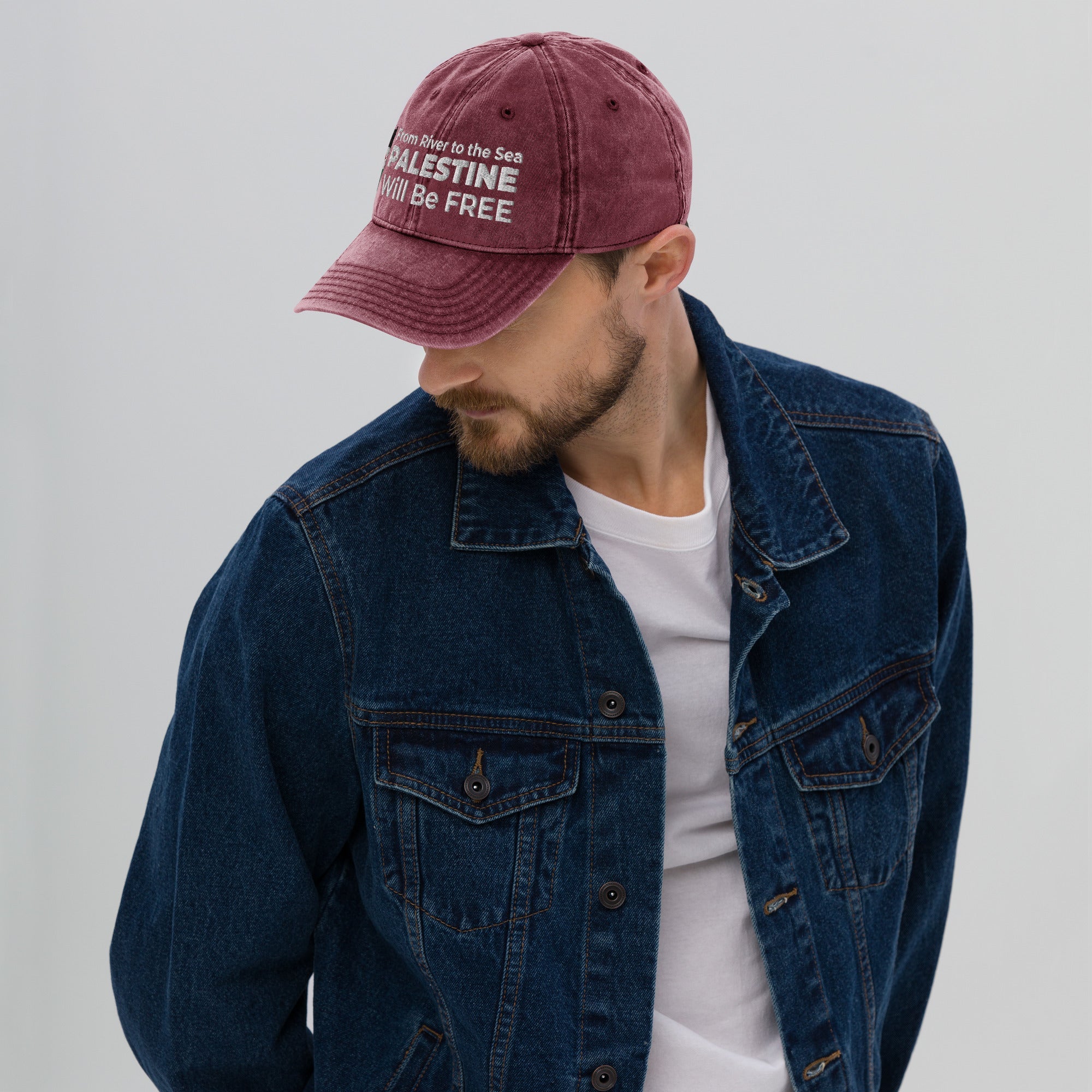 A man wearing a denim jacket and a maroon cap with the text and Palestine flag, on a plain background