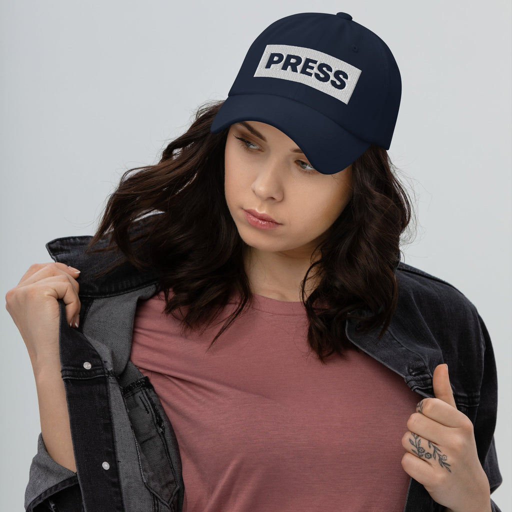 Woman wearing a navy blue cap with 'PRESS' on it against a gray background