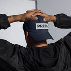 Person wearing a navy blue cap with 'PRESS' on it, held by two hands against a neutral background